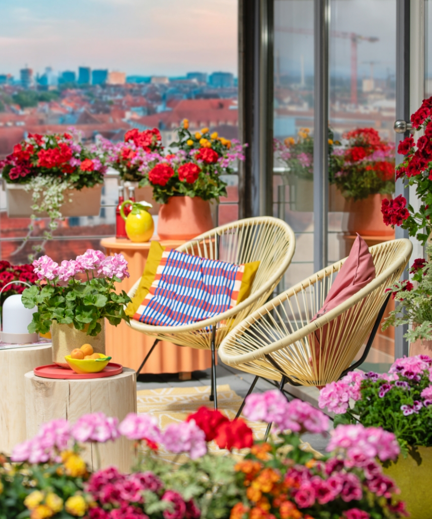 Ein Balkon mit Blick auf die Stadt, voller farbenfroher Geranien in Rot, Rosa und Purpur. Moderne Möbel und bunte Kissen schaffen eine einladende Atmosphäre für entspannte Stunden im Freien.