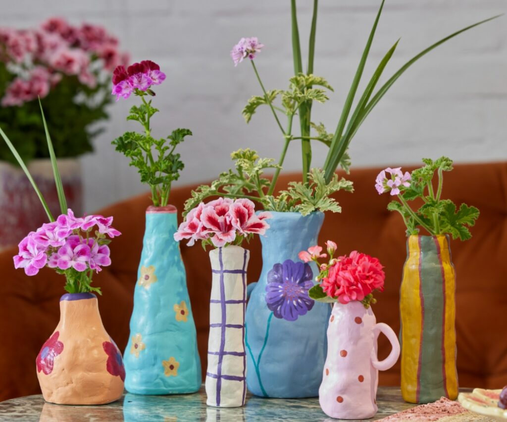 Mehrere kleine, handgeformte Vasen aus Modelliermasse in unterschiedlichen Farben stehen nebeneinander auf einem Tisch und sind jeweils mit geschnittenen Geranien, einzelnen Blumen oder Gräsern bestückt.