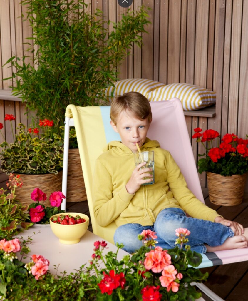 Junge sitzt auf einem Stuhl im Freien und hält ein Glas, umgeben von blühenden Geranien in Töpfen.
