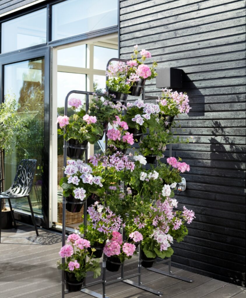 Vertikales Pflanzregal mit rosa und weißen Geranien auf moderner Terrasse, mehrere Töpfe übereinander angeordnet