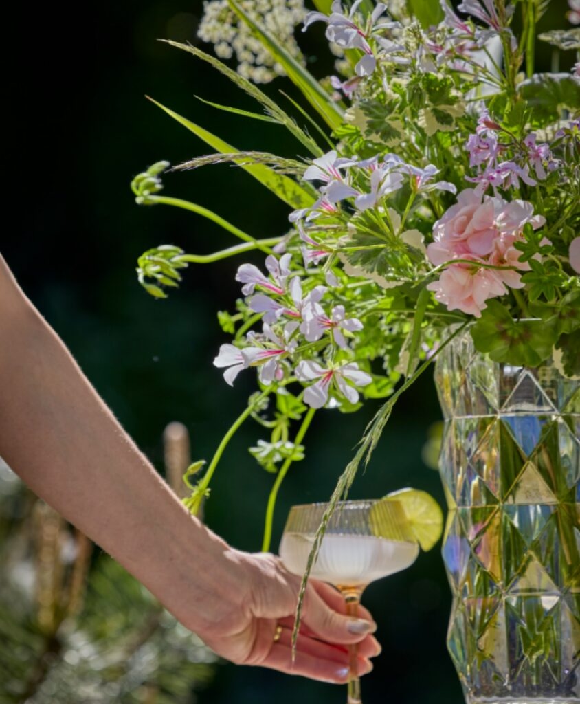 Nahaufnahme eines Blumenstraußes aus Geranien und Gräsern; eine Hand stellt ein Glas mit Getränk neben die Vase.