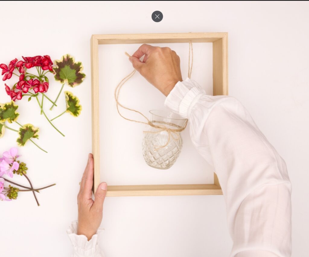 Hände befestigen eine kleine Glasvase mit Juteschnur in einem Holzrahmen; daneben liegen Geranienblüten für eine hängende Blumendeko.