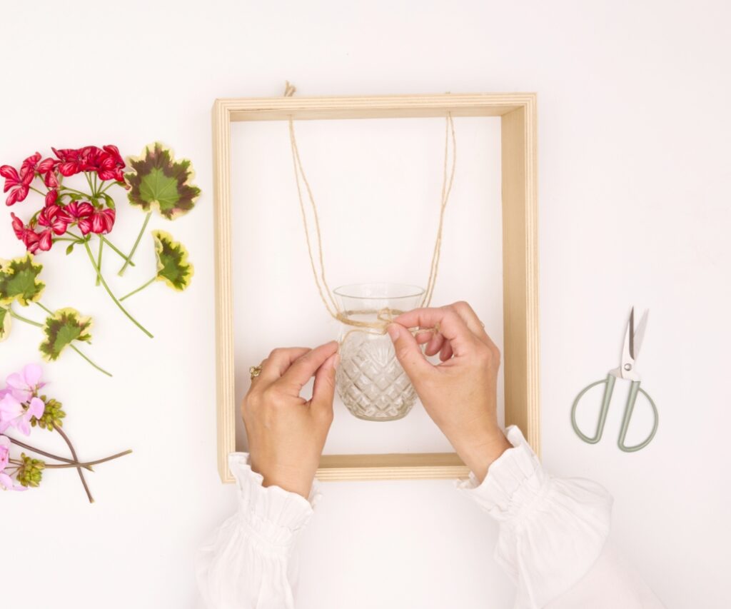 Hände binden Juteschnur um eine Glasvase in einem Holzrahmen; geschnittene Geranien liegen bereit für die Dekoration.