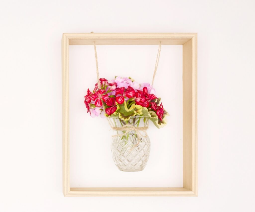 Hängende Glasvase mit rot-rosa Geranien in einem Holzrahmen; die Vase ist mit Juteschnur befestigt.