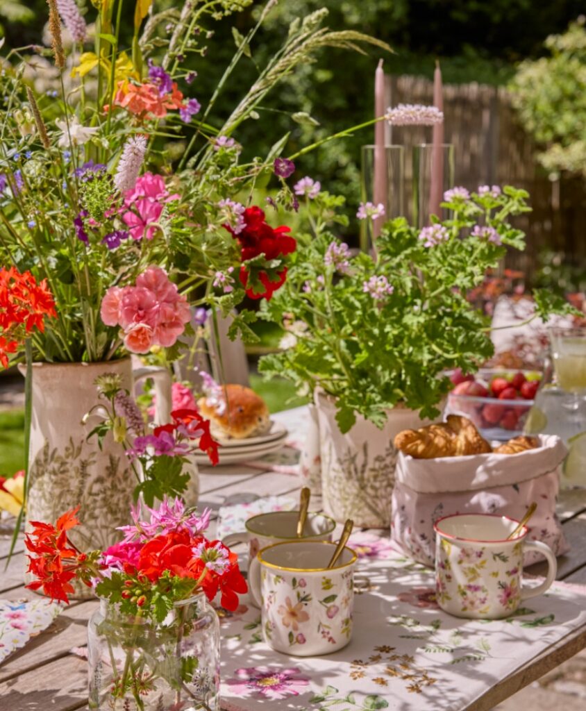 Sommerlich gedeckte Gartentafel mit mehreren Vasen voller Geranien und Wiesenblumen, Geschirr und Frühstücksdetails.