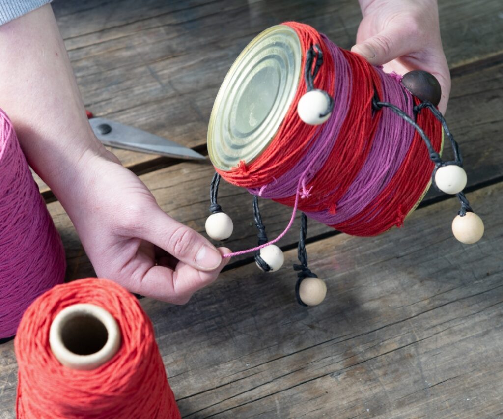 Eine Konservendose wird mit buntem Garn umwickelt, um ein dekoratives Insektenhotel für den Garten mit vielen Geranien zu basteln.