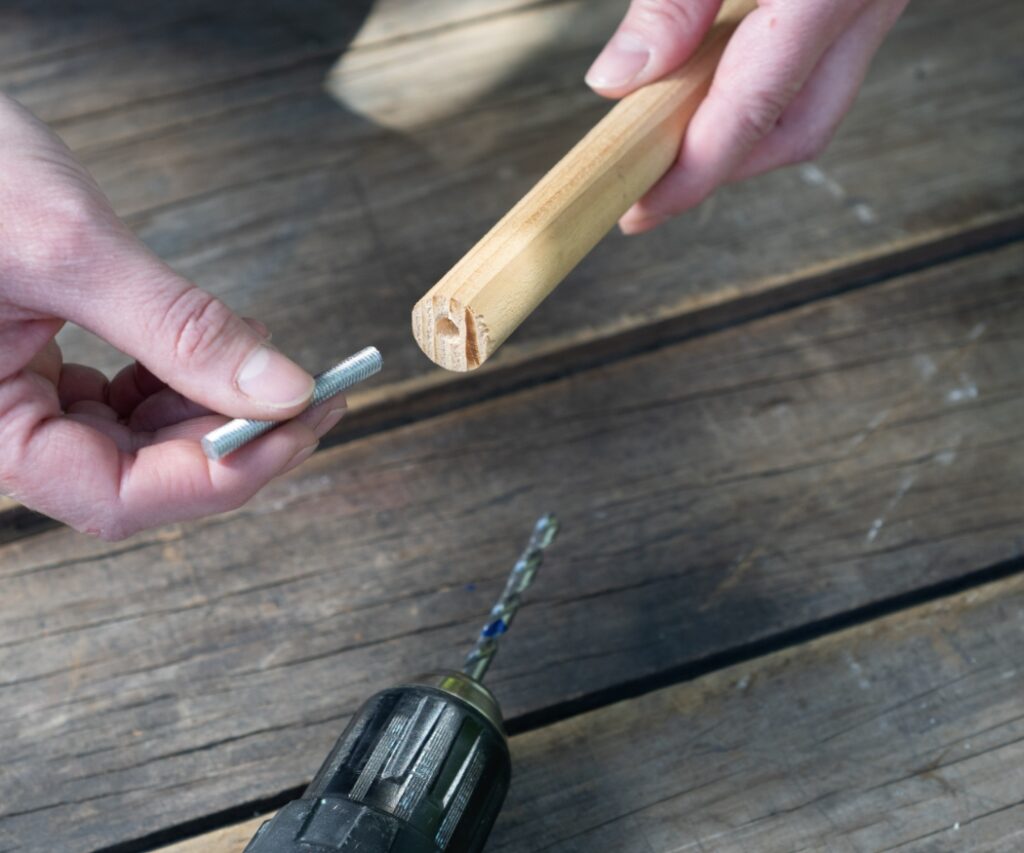 Nahaufnahme der Hände beim Bohren eines Lochs in ein Rundholz und dem Einsetzen einer Gewindestange als stabile Basis für ein DIY-Insektenhotel in der Nähe von Geranien für Bienen.
